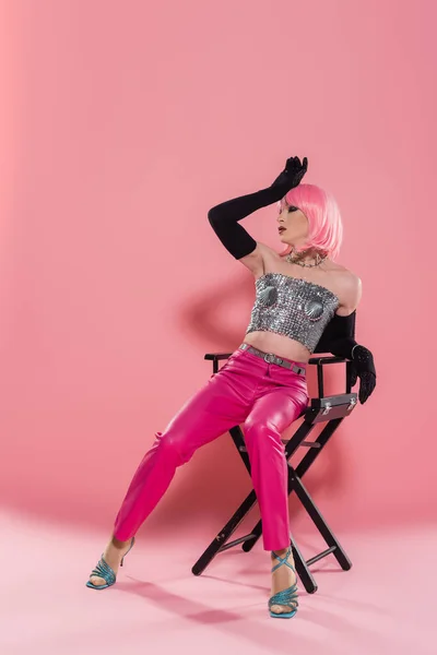 Full length of fashionable drag queen in shiny top posing on chair on pink background — Stock Photo