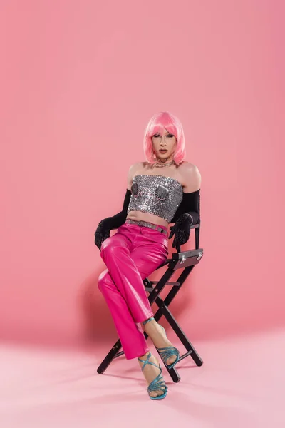 Fashionable drag queen in top and gloves posing while sitting on chair on pink background — Stock Photo