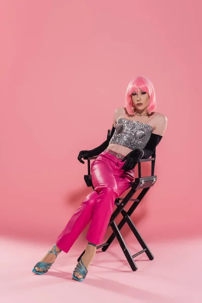 Stylish drag queen in top and wig sitting on chair on pink background — Stock Photo