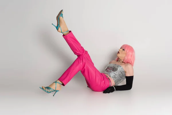 Модный трансгендер в розовом парике и сверкающем топе лежит на сером фоне — стоковое фото