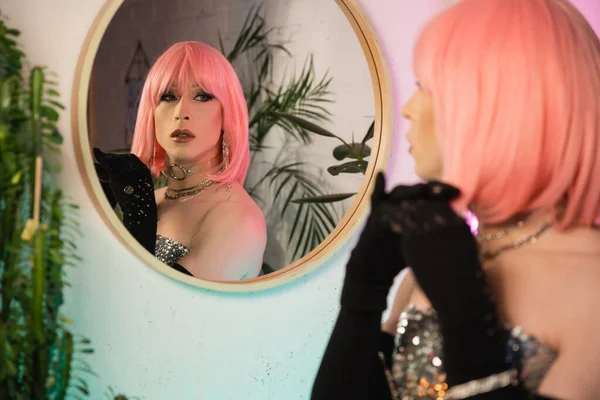 Elegante drag queen en la parte superior y guantes mirando el espejo en casa - foto de stock