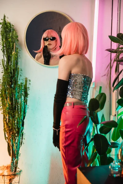 Sfocata drag queen in occhiali da sole in piedi vicino specchio e piante a casa — Foto stock