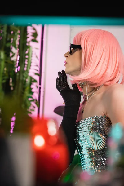 Vista lateral da elegante drag queen em peruca rosa e óculos de sol em pé perto de plantas em casa — Fotografia de Stock