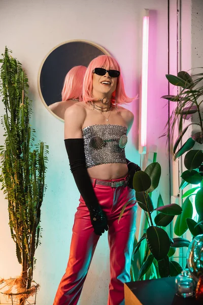 Alegre drag queen en gafas de sol y peluca de pie cerca de las plantas y el espejo en casa - foto de stock