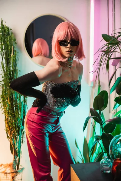 Elegante drag queen en peluca rosa y gafas de sol posando cerca de la iluminación en casa - foto de stock