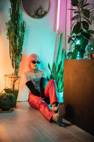 Stylish transgender person in sunglasses sitting on floor near plants at home — Stock Photo