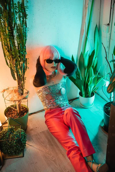 Trendy drag queen in parrucca rosa e occhiali da sole seduti sul pavimento vicino alle piante a casa — Foto stock