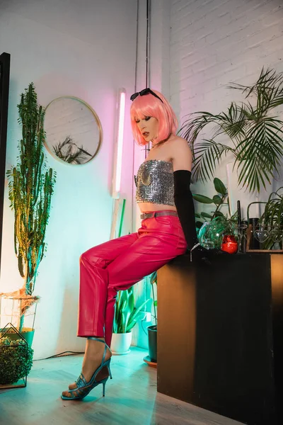Trendy Drag Queen in pinkfarbener Perücke und Sonnenbrille sitzt zu Hause auf einem Tisch neben Pflanzen — Stockfoto