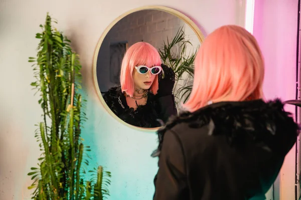 Trendy drag queen en peluca y gafas de sol de pie cerca del espejo y las plantas en casa - foto de stock