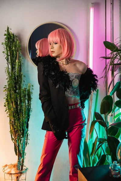 Trendy drag queen en peluca rosa y chaqueta de pie cerca de las plantas en casa - foto de stock