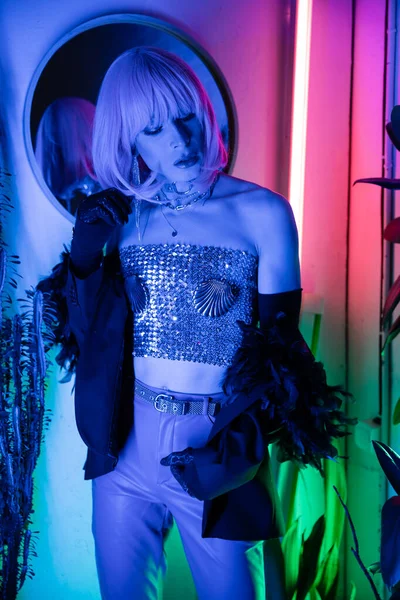 Trendy transgenre personne en perruque et veste debout près de néon lumière à la maison — Photo de stock