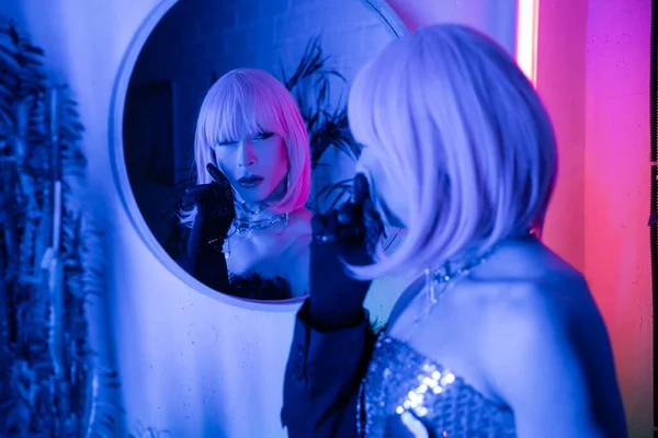 Drag queen à la mode en perruque regardant miroir dans la lumière au néon à la maison — Photo de stock