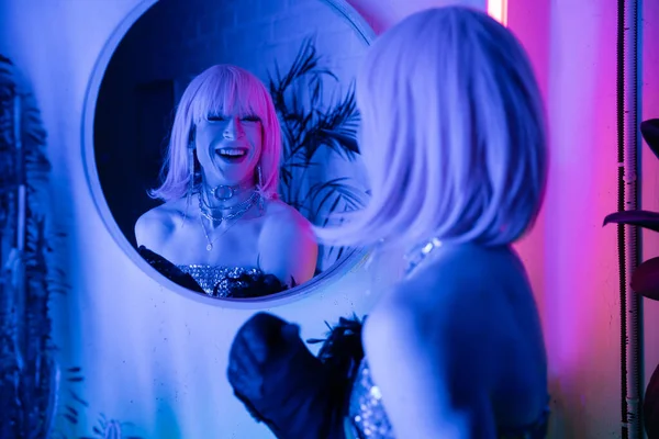 Cheerful drag queen in wig and top looking at mirror in neon light at home — Stock Photo