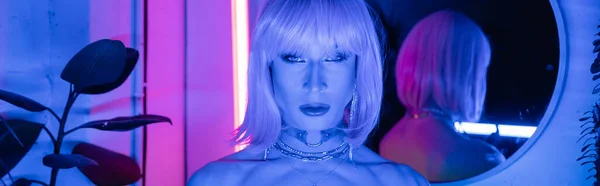 Portrait of drag queen in wig looking at camera in neon lighting at home, banner — Stock Photo