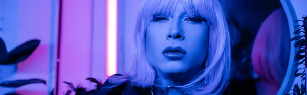 Drag queen em peruca e visagem olhando para a câmera perto de luz de néon em casa, banner — Fotografia de Stock