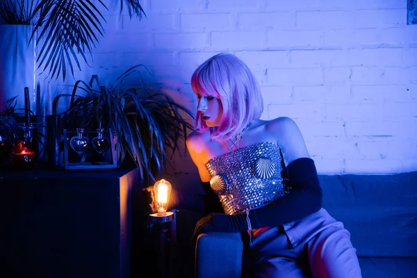 Drag queen in wig and top looking at lamp near plants at home — Stock Photo