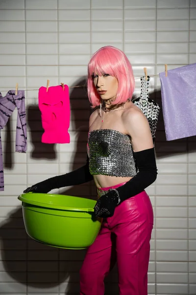 Stylish transgender person in wig and gloves holding basin near clothes in bathroom — Stock Photo