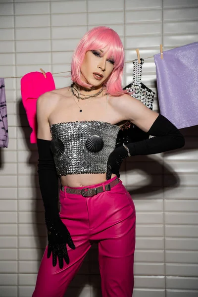 Stylish drag queen in shiny top and gloves looking at camera near clothes on rope in bathroom — Stock Photo
