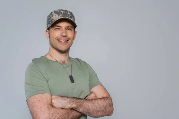 Happy American soldier in t-shirt and military cap standing with folded arms during memorial day isolated on grey — Stock Photo