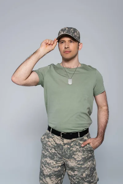 Soldado patriótico en camiseta ajustando gorra militar durante el día conmemorativo aislado en gris - foto de stock