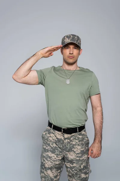 Patriota estadounidense en camiseta y gorra militar saludando durante el día conmemorativo aislado en gris, estandarte - foto de stock