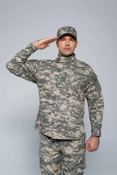 Soldado del ejército patriótico en uniforme militar con gorra saludando durante el día de fiesta conmemorativo aislado en gris - foto de stock
