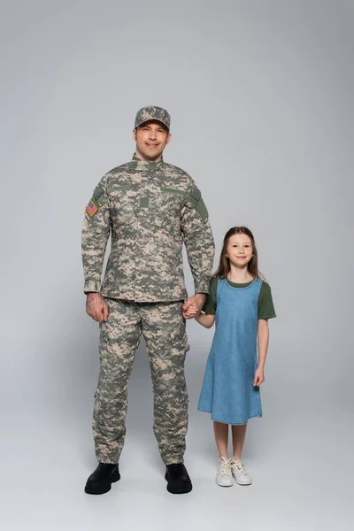 Pleine longueur de soldat de l'armée heureux en uniforme militaire debout avec sa fille pendant la journée commémorative sur gris — Photo de stock