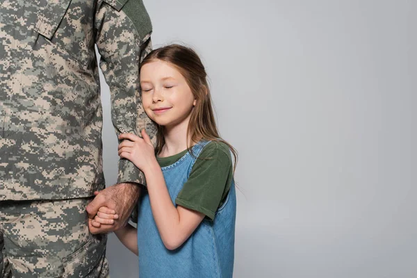 Alegre preadolescente chica abrazando mano de padre en militar uniforme aislado en gris - foto de stock