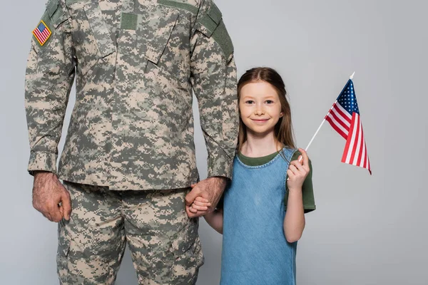 Alegre niña preadolescente sosteniendo la bandera americana y la mano del padre en uniforme militar durante el día conmemorativo aislado en gris - foto de stock