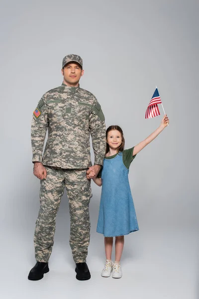 Fröhliches Mädchen mit amerikanischer Flagge und der Hand des Vaters in Militäruniform am Gedenktag auf grau — Stockfoto
