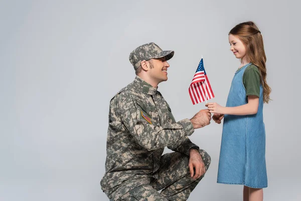 Alegre niña preadolescente dando bandera americana a feliz padre en uniforme militar durante el día conmemorativo aislado en gris - foto de stock