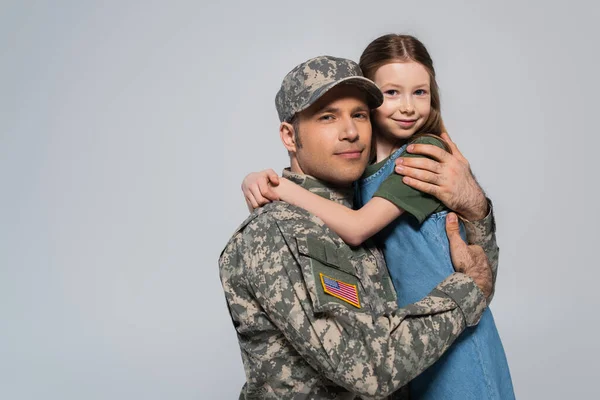 Feliz niña abrazando padre en uniforme militar durante el día conmemorativo aislado en gris - foto de stock