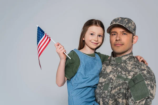 Soldat in Militäruniform hält glückliche Tochter mit amerikanischer Flagge auf grau — Stockfoto