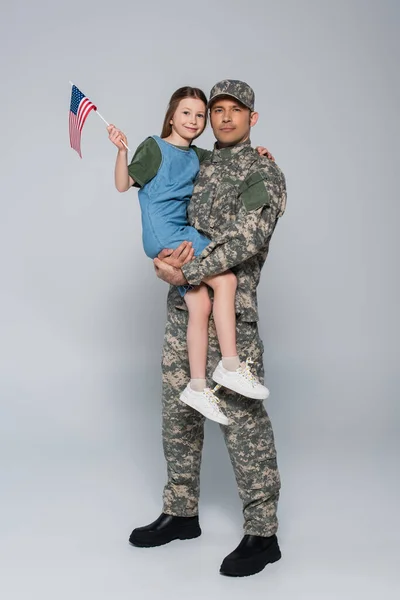 Militaire en uniforme militaire tenant dans les bras fille joyeuse avec drapeau américain sur gris — Photo de stock