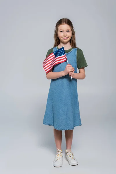 Longitud completa de niña preadolescente feliz y patriótica sosteniendo la bandera de América durante el día conmemorativo en gris - foto de stock