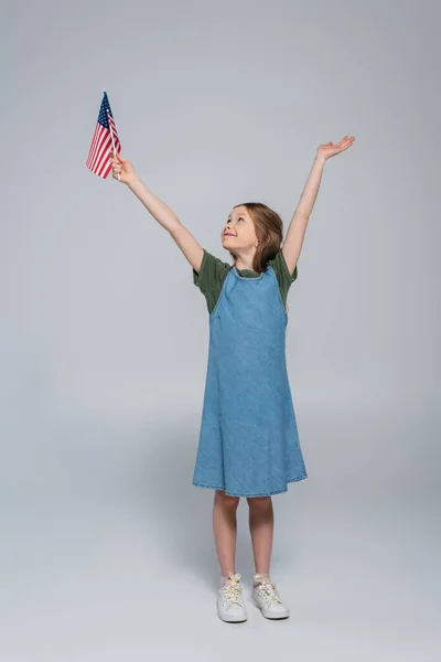 Full length of patriotic preteen girl smiling while holding flag of United States of America during memorial day on grey — Stock Photo