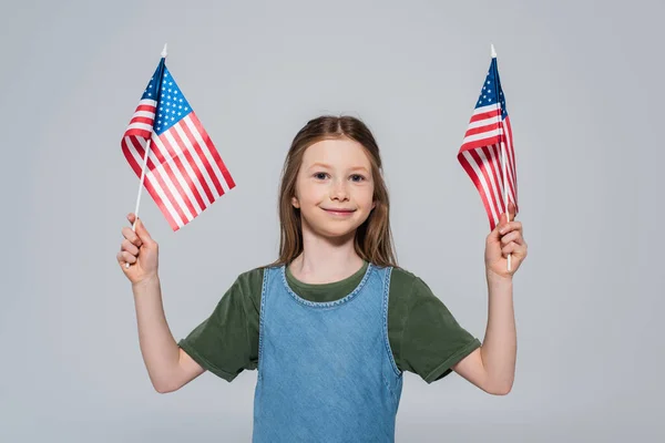 Niña preadolescente patriótica sonriendo mientras sostiene banderas de América durante el día conmemorativo aislado en gris - foto de stock