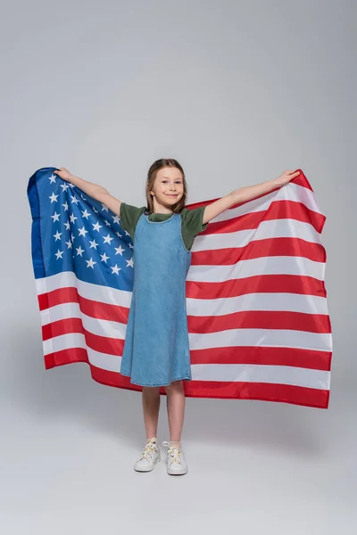 Longitud completa de la niña preadolescente patriótica sonriendo mientras sostiene la enorme bandera de Estados Unidos durante el día memorial en gris - foto de stock