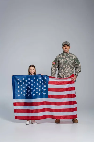 Serviceman in military uniform holding flag of United States with happy daughter on grey — Stock Photo