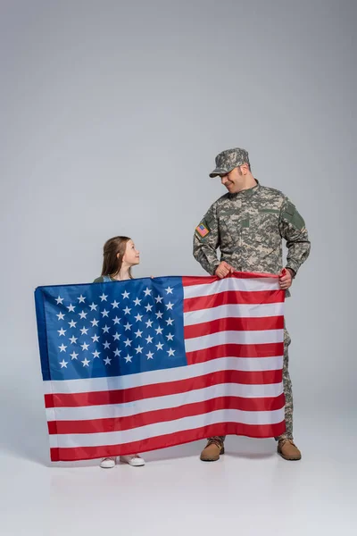 Cheerful serviceman in military uniform holding flag of United States with happy daughter on grey — Stock Photo