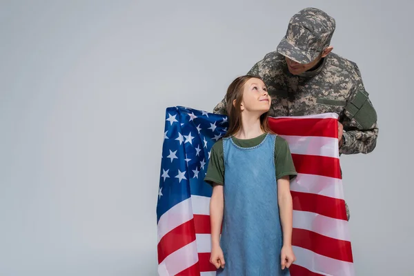 Militar en uniforme militar sosteniendo bandera de Estados Unidos cerca hija feliz aislado en gris - foto de stock