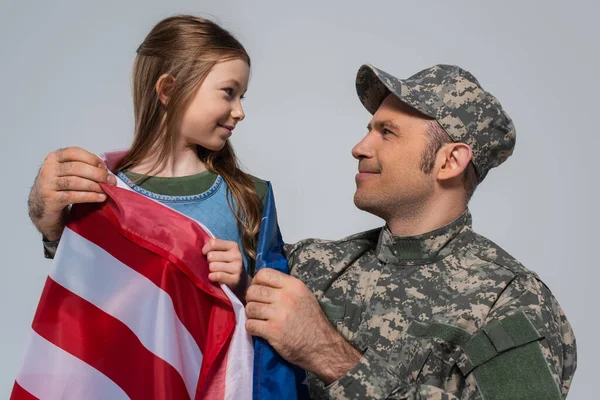 Militaire en uniforme militaire regardant fille heureuse enveloppée dans le drapeau des États-Unis d'Amérique isolé sur gris — Photo de stock