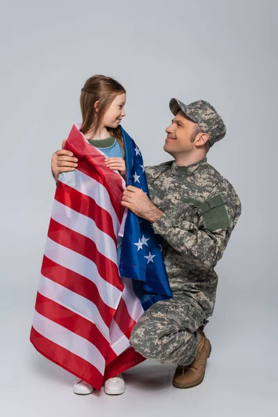 Feliz soldado de uniforme militar mirando a su hija envuelta en la bandera de los Estados Unidos de América en gris - foto de stock