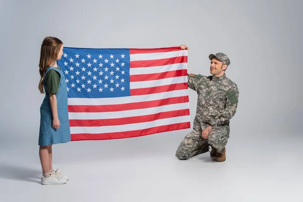 Happy serviceman in military uniform holding flag of USA with daughter on grey — Stock Photo