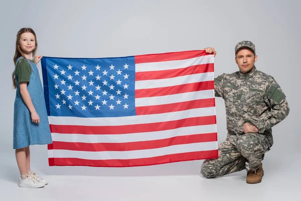 Serviceman in military uniform holding flag of USA with cheerful daughter on grey — Stock Photo