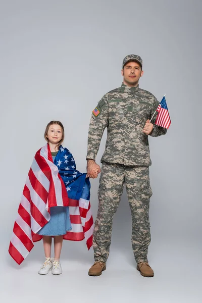 Serviceman in military uniform holding flag of USA and hand of happy daughter on grey — Stock Photo