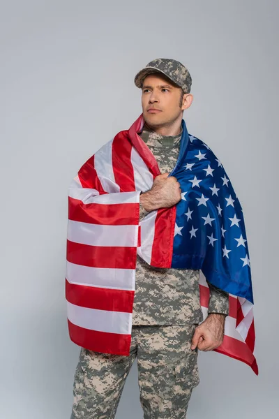 Soldado del ejército patriótico en uniforme de camuflaje envuelto en la bandera de los Estados Unidos de América durante el día conmemorativo aislado en gris - foto de stock