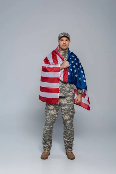 Toute la longueur du soldat de l'armée patriotique en uniforme de camouflage enveloppé dans le drapeau des États-Unis d'Amérique pendant la journée commémorative sur gris — Photo de stock