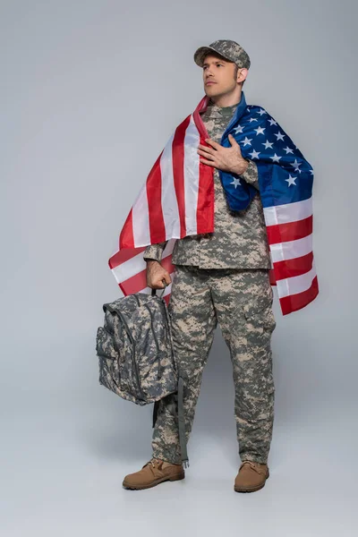 Soldado del ejército patriótico en uniforme de camuflaje envuelto en la bandera de Estados Unidos de América de pie con mochila en gris - foto de stock