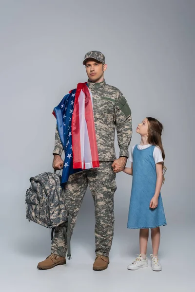 Soldado del ejército en uniforme con bandera de los Estados Unidos de América sosteniendo la mochila y de pie con su hija en gris - foto de stock